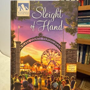 Peut inclure: Couverture du livre "Sleight of Hand" de Carole Jefferson. L'illustration montre une fête foraine au coucher du soleil, avec une grande roue, une foule et le texte "Blake County Fairgrounds". Fait partie de la série "Mysteries of Silver Peak".