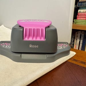 Puede incluir: Un troquel de manualidades gris y rosa con la palabra "Rose" impresa en el lateral. El troquel tiene una parte superior rosa y un diseño floral decorativo en la base. El troquel está sobre una superficie blanca.