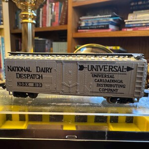 May include: A model train car with the text "National Dairy Despatch" and "Universal Carloadings & Distributing Company" printed on the side. The car is light gray with black wheels and is displayed on a clear surface.