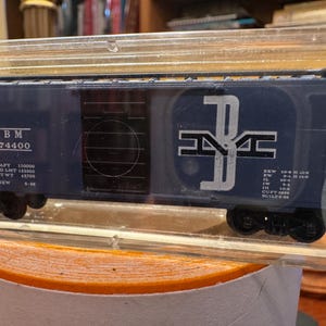 May include: A blue model train car with black wheels and white lettering. The car has the letters "BM" and the number "74400" printed on the side. The car is in a clear plastic case.