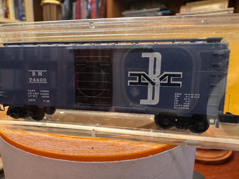 May include: A blue model train car with black wheels and white lettering. The car has the letters "BM" and the number "74400" printed on the side. The car is in a clear plastic case.