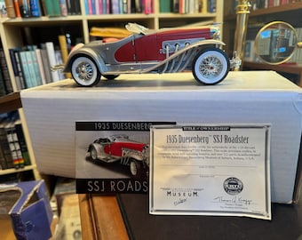 May include: A 1:18 scale die-cast model of a 1935 Duesenberg SSJ Roadster in red and silver. The car features wire-spoke wheels with white-wall tires and external exhaust pipes. It includes a certificate of authenticity and a product box.