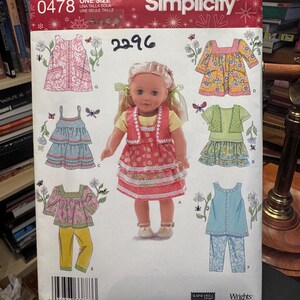 Puede incluir: Un patrón de costura Simplicity para ropa de muñecas, con varios diseños de atuendos en rosa, azul, amarillo y verde. El patrón incluye una muñeca con un vestido rojo. El paquete tiene el número 2296 y el texto "ONE SIZE".