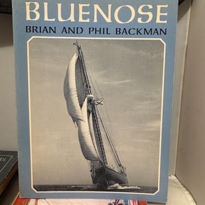 May include: A book titled "Bluenose" by Brian and Phil Backman. The cover features a black and white image of a sailboat with billowing sails on the water. The book has a light blue border and the title is in large, bold letters.