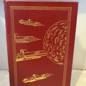 Puede incluir: Un libro encuadernado en cuero rojo con un diseño dorado de naves espaciales y una luna en la portada.