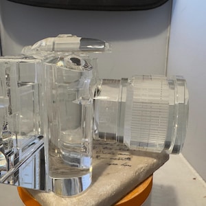 May include: A clear, glass camera sculpture with a lens and flash. The camera is displayed on a decorative base. The sculpture is made of clear glass and has a detailed design. The camera is a decorative item.