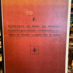 May include: A rectangular book cover with a red gradient background featuring the text F. Elastolin-Figures and Wooden Toys Manufactures, Produits de Figures d'Elastolin et de Bimbeloterie en bois, Figuras de Elastolin y juguetes finos de madera.