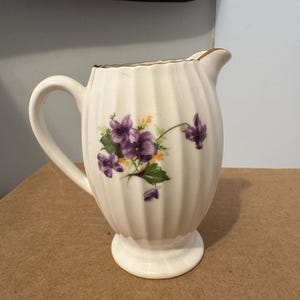 Puede incluir: Una crema de cerámica blanca con un diseño acanalado y un motivo floral de violetas moradas y hojas verdes. La crema tiene un borde dorado y un asa. La crema está sobre una superficie marrón.