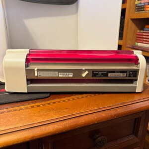 Puede incluir: Una máquina Xyron Wishblade blanca y gris con una parte superior roja. La máquina está sobre una mesa de madera. El texto "Xyron Wishblade" y "H701803452" son visibles en la máquina.
