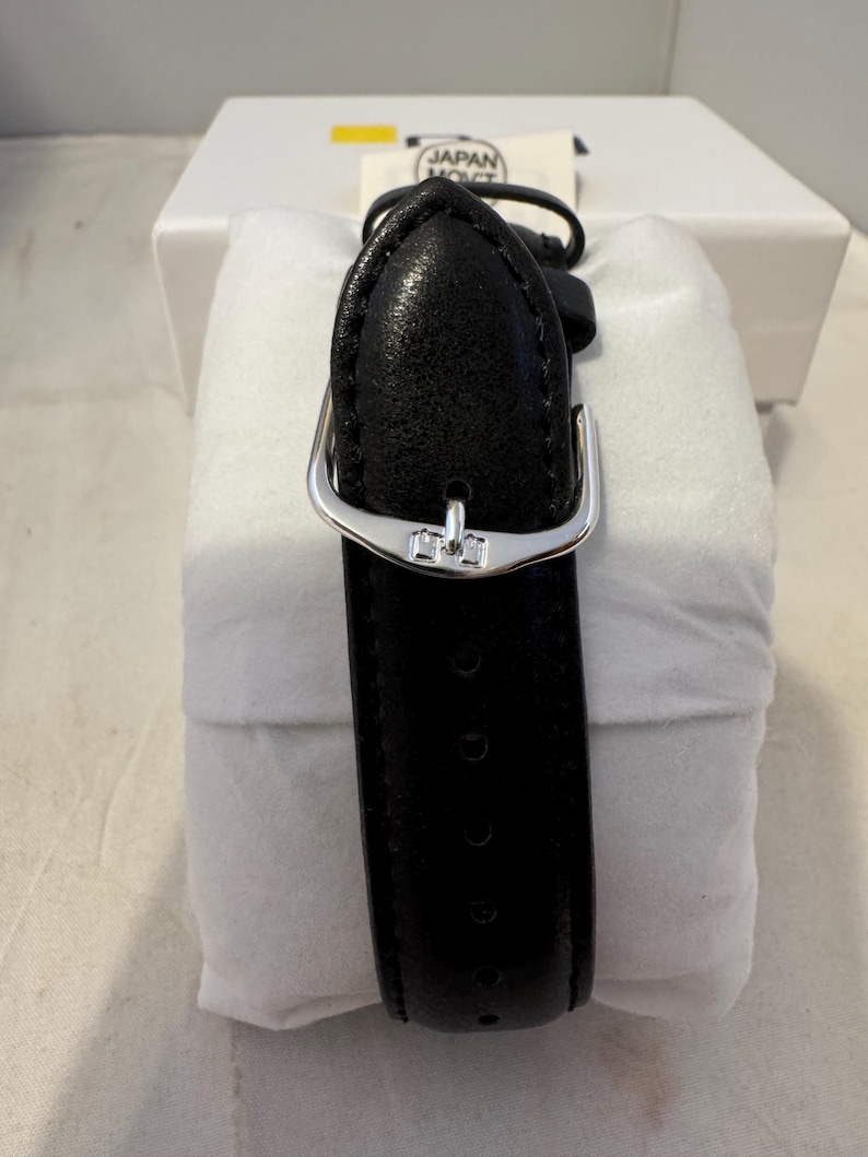 May include: Black leather watch band with a silver buckle. The band is in a white box with the text "JAPAN MOV'T" on a label.