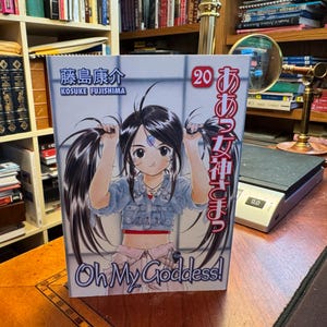 May include: A manga volume titled Oh My Goddess! number 20 by Kosuke Fujishima. The cover features a black-haired character with twin tails wearing a light blue denim jacket and a pink skirt, with Japanese text and the number 20 printed in red.