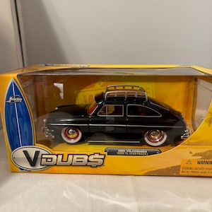 May include: A black 1965 Volkswagen 1600 TL Fastback model car in a clear display case with a yellow and black box. The box has the words "V DUBS" on it. The car has a roof rack and whitewall tires.