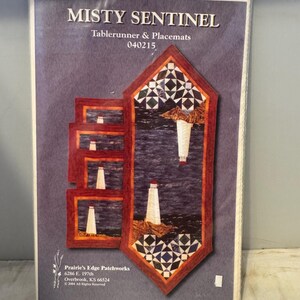 May include: A quilting pattern for a table runner and placemats featuring a lighthouse scene with a blue and white colour scheme. The pattern is titled "Misty Sentinel" and is from Prairie's Edge Patchworks.