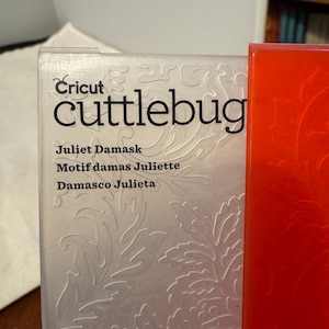 以下が含まれることがあります： 「Cricut Cuttlebug」の箱のクローズアップ。前面には「Cuttlebug」と「Juliet Damask」の文字が印刷されています。箱は白で、エンボス加工された花の模様があります。右側には赤い箱が部分的に見えています。