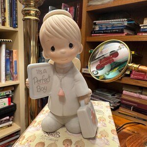 May include: A ceramic figurine of an angel boy holding a sign that reads "It's A Perfect Boy." The angel is wearing a white robe and has a halo. The figurine also holds a small suitcase. The figurine is sitting on a decorative box.