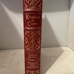 May include: A red leather-bound book with gold lettering. The title is "Kingdom of Cages" by Sarah Zettel. The book is a signed first edition.