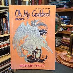 May include: A softcover manga book titled Oh My Goddess! Mystery Child by Kosuke Fujishima. The orange cover features a central illustration of a winged figure with long hair and two smaller character portraits below, all rendered in a manga art style.
