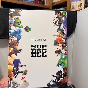 Puede incluir: Un libro de tapa dura titulado "THE ART OF SUPERCELL" con coloridos personajes de dibujos animados. El libro está en un estuche negro. La portada incluye varios personajes en poses de acción, con un fondo blanco.
