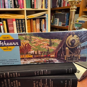 May include: A rectangular Athearn Trains in Miniature box featuring a detailed illustration of a steam locomotive and passenger cars in a mountainous landscape. The blue and yellow packaging includes the text Authentic-Realistic and Made in U.S.A.