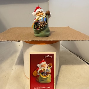 Könnte beinhalten: Hallmark Weihnachtsschmuck mit dem Weihnachtsmann, der einen grünen Sack voller Spielzeug hält. Der Schmuck hat einen rot-weißen Weihnachtsmannhut und ein lächelndes Gesicht. Eine Karte mit dem Text "Santa's Magic Sack" ist ebenfalls sichtbar.