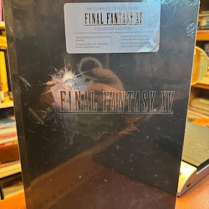 May include: A black Final Fantasy XV Collector's Edition hardcover strategy guide wrapped in clear plastic. The cover features the game title in silver text and a sticker detailing contents like a 32-page art gallery and a 34 by 22 inch map.