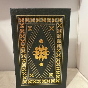 Puede incluir: Un libro de tapa dura de color verde oscuro con un diseño en relieve dorado en la portada. El diseño presenta un patrón geométrico con un motivo central de una flor estilizada.