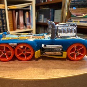 May include: A blue toy truck with a chrome engine block and dual exhaust pipes. The vehicle features six bright orange wheels, yellow side accents, and a metallic silver fuel tank. The body is decorated with yellow and white graphic patterns.