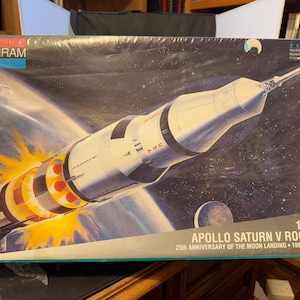 Può includere: Scatola del kit di modelli Monogram con un'illustrazione del razzo Apollo Saturn V nello spazio. L'illustrazione mostra il razzo che decolla con fiamme e la scritta "USA". La scatola include anche la scritta "25th Anniversary of the Moon Landing 1969 to 1994".