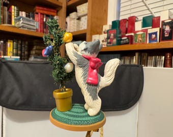 Könnte beinhalten: Eine Keramikfigur einer Katze mit rotem Schal, die nach einem kleinen Weihnachtsbaum greift. Der Baum ist mit bunten Ornamenten geschmückt und steht in einem goldenen Topf. Die Figur steht auf einem kleinen, runden Tisch.