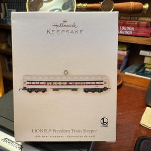 May include: A Hallmark Keepsake Christmas ornament featuring a Lionel Freedom Train Sleeper car. The train car is white with red and black accents, and the word "FREEDOM" is printed on the side. The box is white with the Hallmark and Lionel logos.