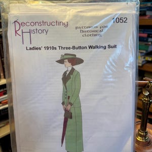 May include: A sewing pattern envelope labeled Reconstructing History 1052 Ladies' 1910s Three-Button Walking Suit. The cover features an illustration of a person wearing a green suit with a dark collar, a wide-brimmed hat, and holding a dark umbrella.