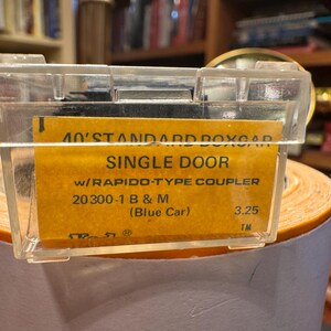 May include: A clear plastic case containing a yellow label with black text. The label reads "40' STANDARD BOXCAR SINGLE DOOR" and other details about a model train car. The case is resting on a roll of orange paper.