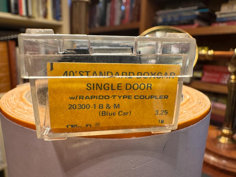 May include: A clear plastic case containing a yellow label with black text. The label reads "40' STANDARD BOXCAR SINGLE DOOR" and other details about a model train car. The case is resting on a roll of orange paper.