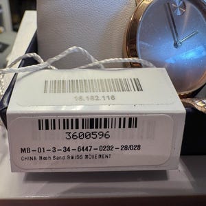 May include: A gold-toned wristwatch with a silver face and a mesh band. The watch is displayed with its original tag, which includes a barcode and the number 3600596. The tag also states "CHINA Mesh Band SWISS MOVEMENT".