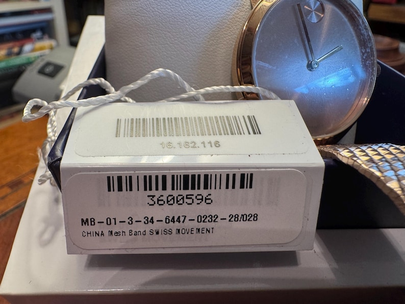 May include: A gold-toned wristwatch with a silver face and a mesh band. The watch is displayed with its original tag, which includes a barcode and the number 3600596. The tag also states "CHINA Mesh Band SWISS MOVEMENT".