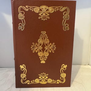 May include: A brown leather-bound book with gold-colored decorative designs on the cover. The designs are floral and intricate.