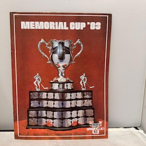 May include: A vintage poster with the text "MEMORIAL CUP '83" at the top. The poster features a large silver trophy, two hockey player figurines, and a tiered base. The background is a deep red.