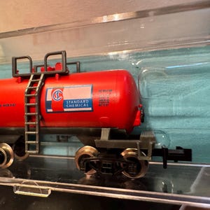 May include: A red model train tank car with the text "STANDARD CHEMICAL" and a ladder. The model train car is on a track and has several wheels. The model train car is in a clear plastic case.