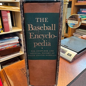 Könnte beinhalten: Ein gebundenes Buch mit dem Titel The Baseball Encyclopedia, The Complete and Official Record of Major League Baseball. Das Buch hat einen dunkelgrünen Mittelteil mit goldener Schrift, flankiert von strukturierten braunen Stoffeinbänden, alles in einem abgenutzten, dunklen, marmorierten Schuber.