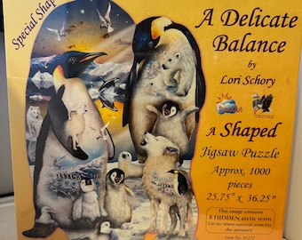 May include: A 1000-piece jigsaw puzzle featuring a delicate balance of penguins and polar bears on a snowy landscape. The puzzle is shaped like a penguin and measures 25.75 inches by 36.25 inches. The box features the title "A Delicate Balance" by Lori Schory.