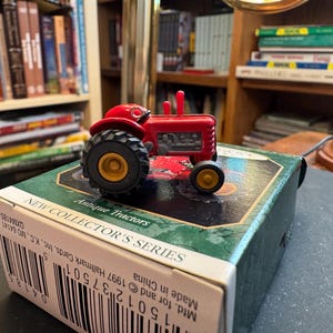 May include: A red toy tractor with black tires and yellow rims sits atop a green and white box. The box reads "NEW COLLECTOR'S SERIES" and "Antique Tractors". The tractor is a miniature replica.