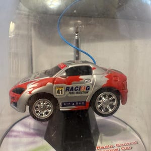 Könnte beinhalten: Ein kleines, silber-rotes ferngesteuertes Spielzeugauto mit dem Wort "RACING" und der Nummer "41" auf der Seite. Das Auto hat eine blaue Antenne und wird in einem transparenten Kunststoffbehälter präsentiert.