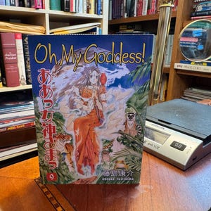May include: A manga volume titled Oh My Goddess! volume 9 by Kosuke Fujishima. The cover features a central female character in an orange dress surrounded by a tiger, a lemur, and lush foliage. The text includes the English and Japanese titles.