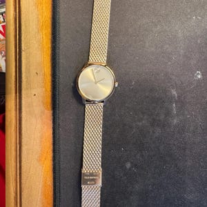 May include: Gold-toned wristwatch with a mesh band. The watch face is round with a minimalist design, featuring a single dot at the 12 o'clock position. The band is made of a fine, woven metal. The word "BOLD" is printed on the top of the band.
