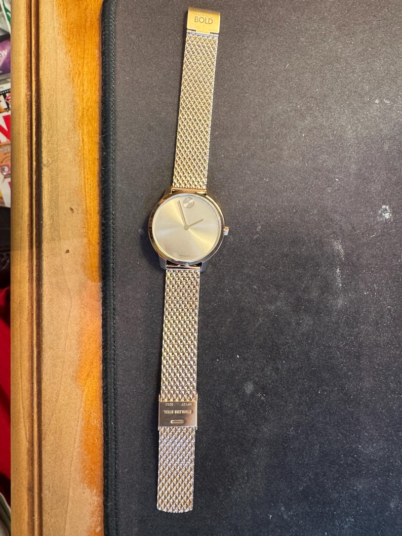May include: Gold-toned wristwatch with a mesh band. The watch face is round with a minimalist design, featuring a single dot at the 12 o'clock position. The band is made of a fine, woven metal. The word "BOLD" is printed on the top of the band.