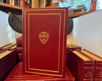 May include: A stack of books with red covers and gold lettering. The front book has a gold-framed border and a gold emblem. The books are part of "The Harvard Classics" collection.