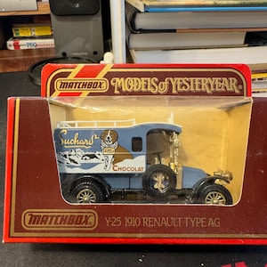 May include: A blue Matchbox Models of Yesteryear die-cast toy car, a 1910 Renault Type AG, with a Suchard chocolate advertisement on the side. The car has a dog on the side of the box and the text "CHOCOLAT" on the side of the car.