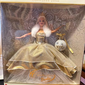 May include: A gold and white Barbie doll wearing a white fur-trimmed gown and a crown. The doll is holding a gold and white ornament with the year 2000 on it. The box says "Special 2000 Edition" and "Celebration Barbie".