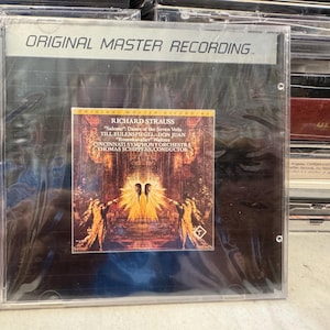 Puede incluir: Un CD con el texto "ORIGINAL MASTER RECORDING" en la parte superior. La portada presenta un álbum de Richard Strauss con la Orquesta Sinfónica de Cincinnati. La carátula muestra dos figuras con una luz brillante.
