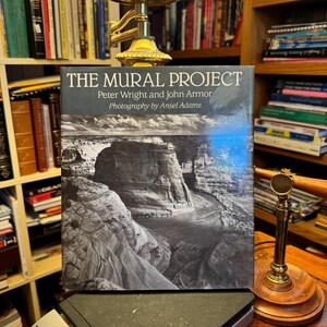May include: A hardcover book titled The Mural Project by Peter Wright and John Armor with photography by Ansel Adams. The cover features a black and white landscape photograph of a river winding through a canyon under a cloudy sky.